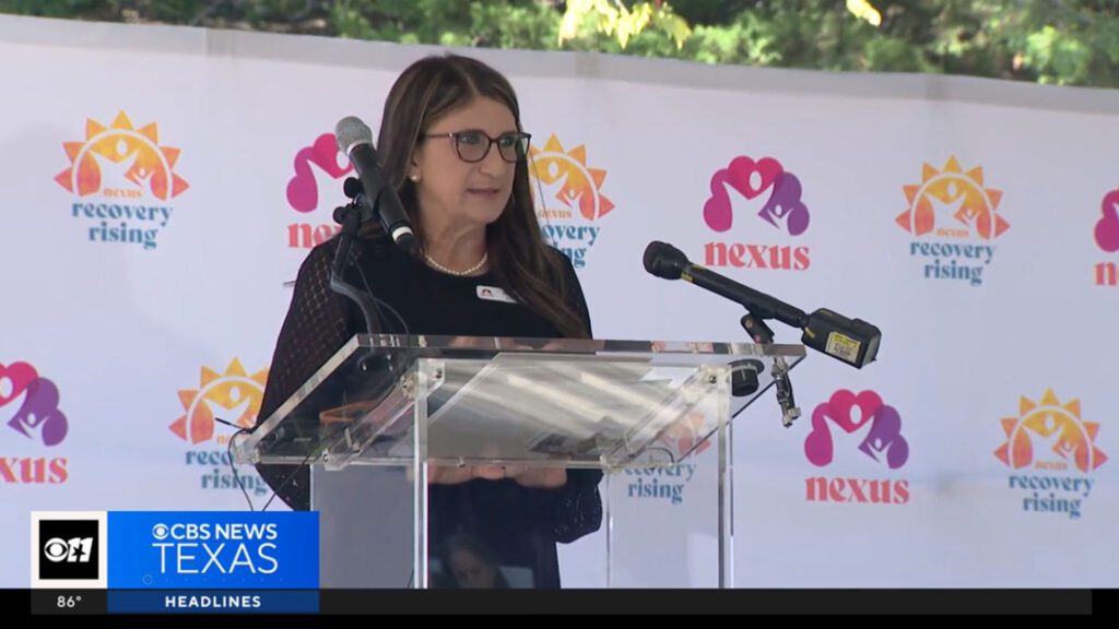 Woman speaking at a podium in front of a Nexus Recovery Rising banner