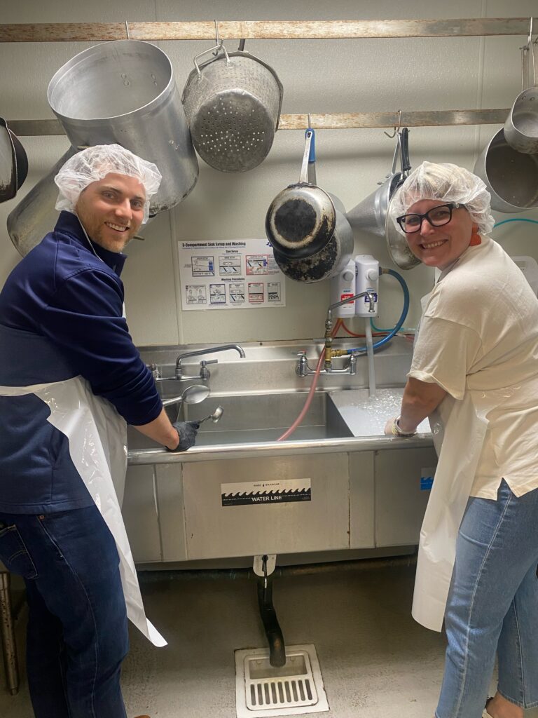 two volunteers wearing protective hair nets washing dishes in industrial sink smiling and front facing with pots and pans hanging on wall in background